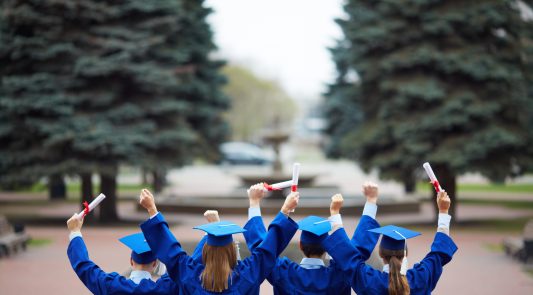 Backs of ecstatic students in graduation gowns holding diplomas Backs of ecstatic students in graduation gowns holding diplomas