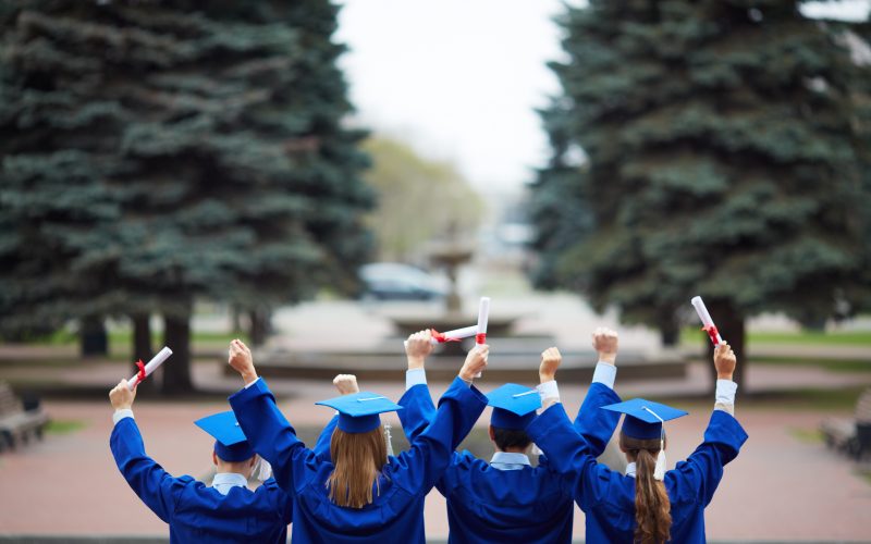 Backs of ecstatic students in graduation gowns holding diplomas Backs of ecstatic students in graduation gowns holding diplomas